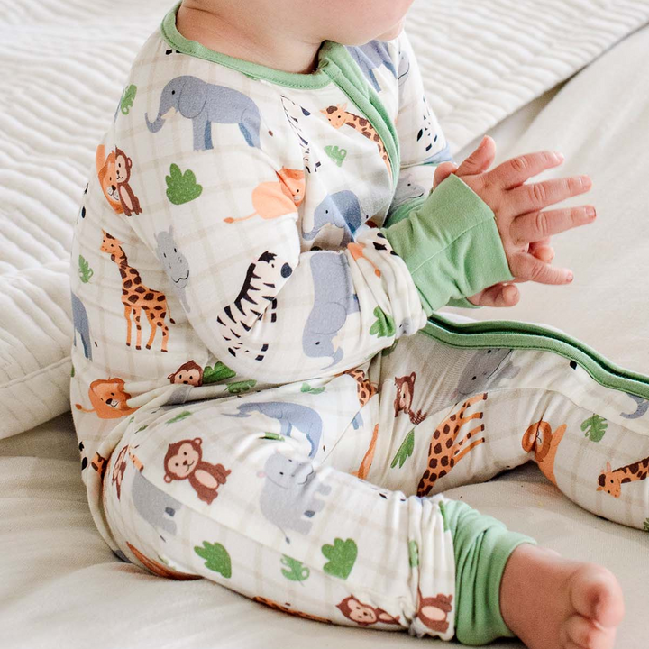Close-up of a seated baby with clasped hands, wearing a Caden Lane cream bamboo convertible zipper romper in a Wild Child print featuring elephants, giraffes, lions, and monkeys.