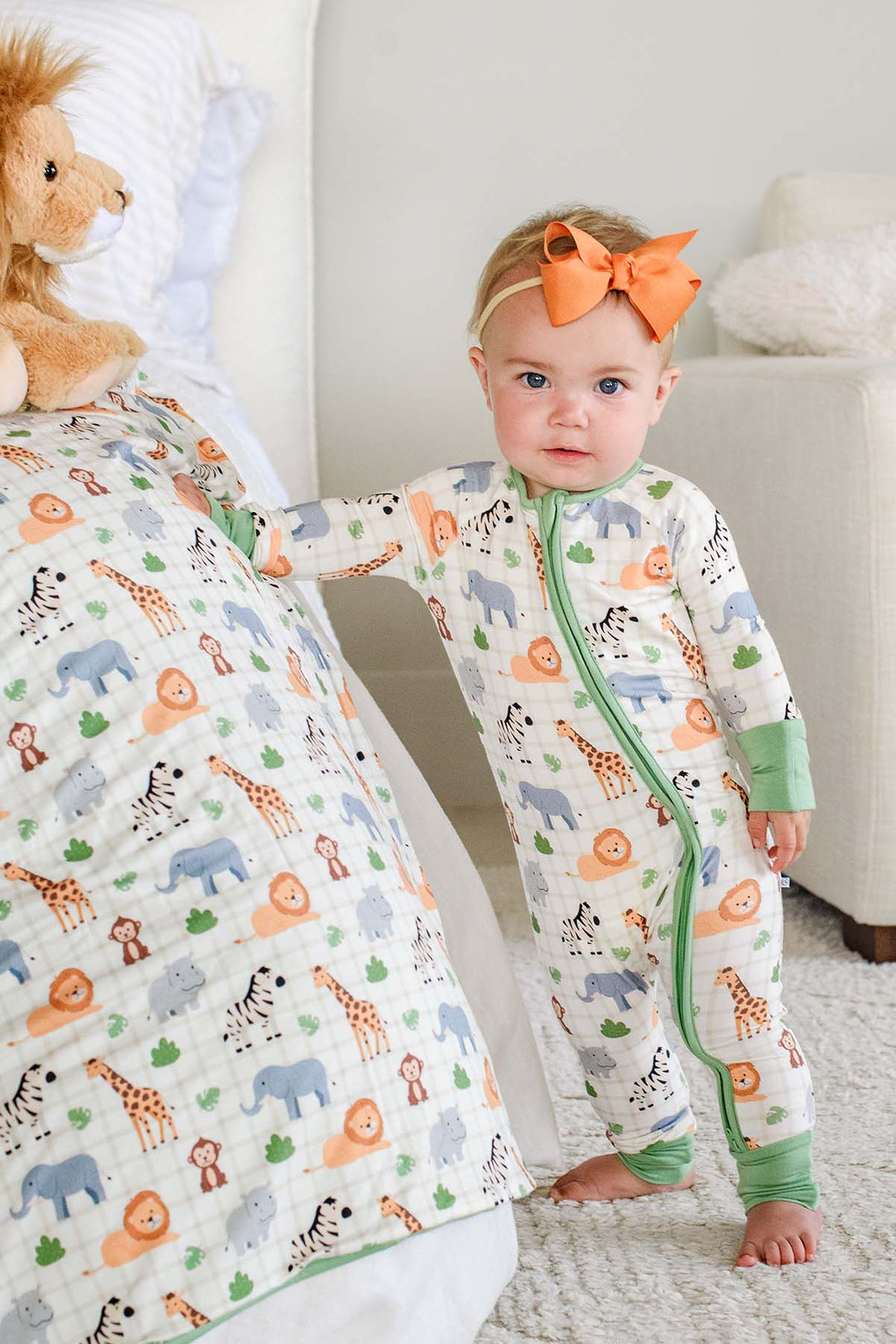 Baby girl standing beside a bed in a Caden Lane convertible zip romper in a Wild Child print, next to a matching blanket and lion plush, paired with an orange bow headband.