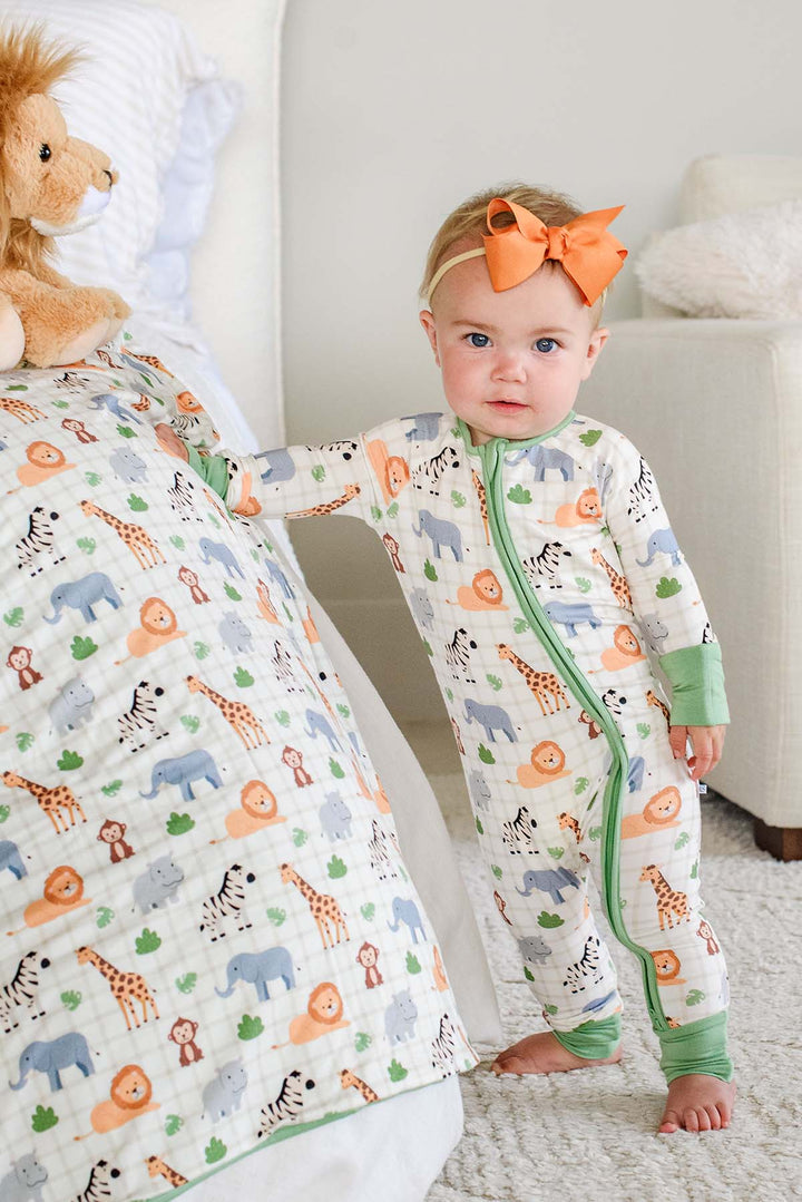Baby girl standing beside a bed in a Caden Lane convertible zip romper in a Wild Child print, next to a matching blanket and lion plush, paired with an orange bow headband.