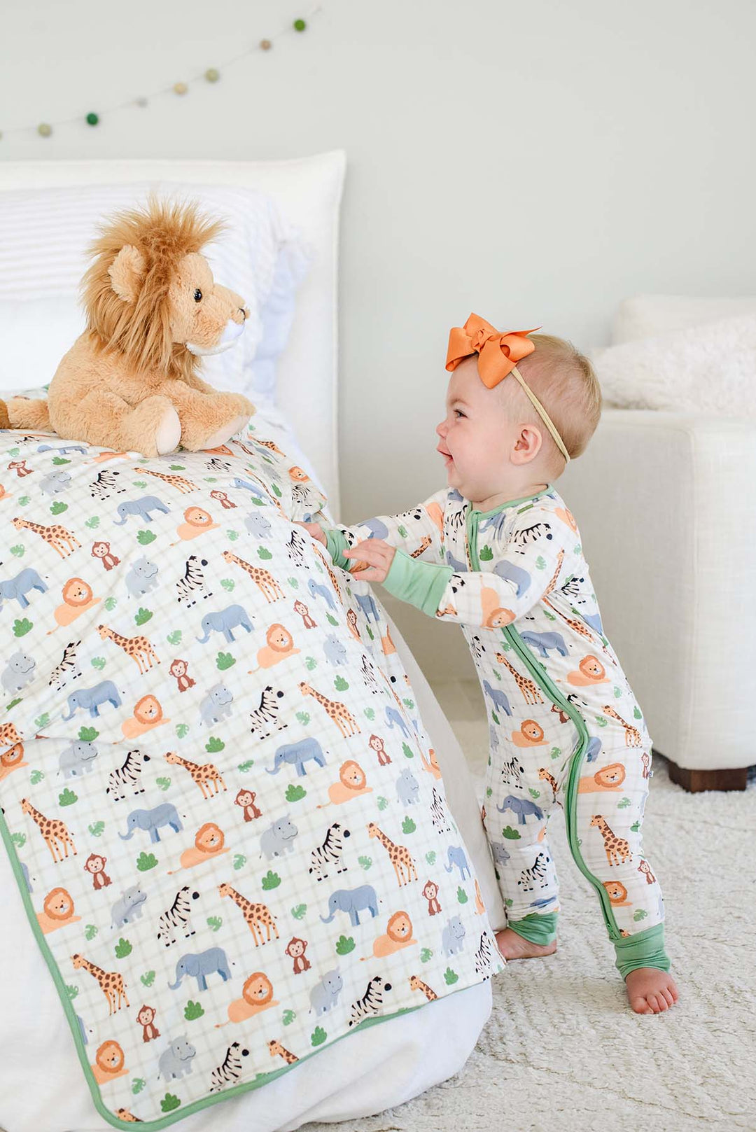 Laughing baby girl standing at the edge of a bed, reaching for a Caden Lane Wild Child bamboo blanket, paired with a matching zip romper, an orange bow and a lion plush.