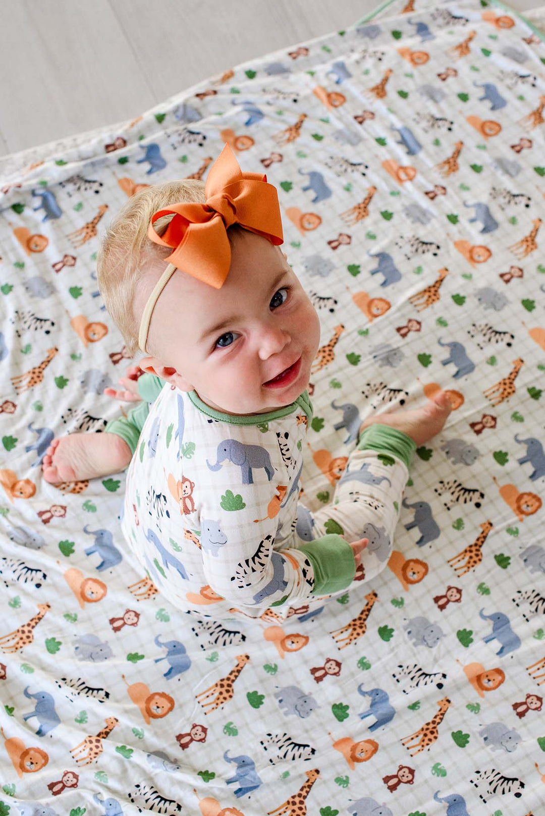 Overhead view of a smiling baby girl looking up while sitting on a Caden Lane Wild Child bamboo blanket, paired with a matching romper and an orange bow headband.