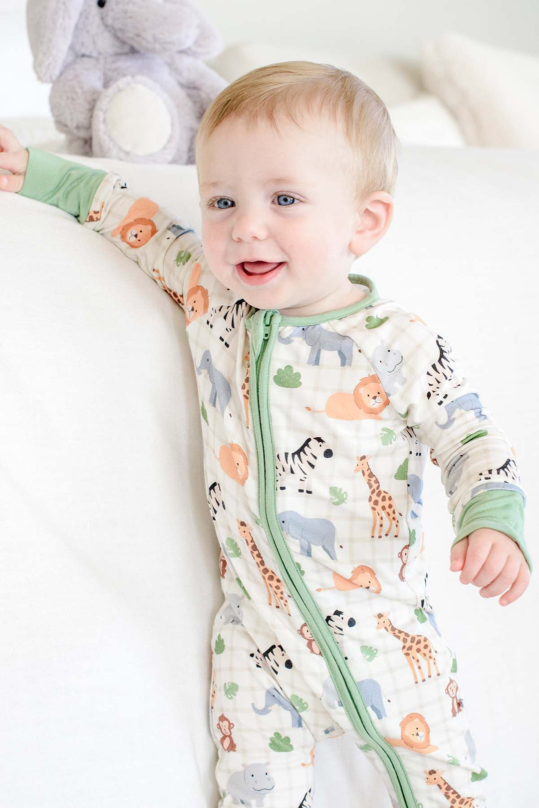 Laughing baby boy standing against a bed in a Caden Lane convertible zip romper in a Wild Child print featuring lions, zebras and giraffes, with an elephant plush behind him.