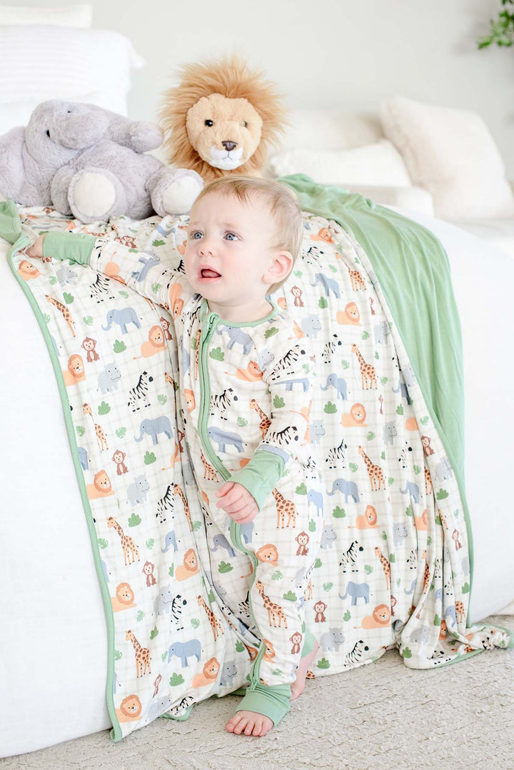 Baby boy standing against a bed draped in a Caden Lane Wild Child bamboo blanket with a sage green reverse, paired with a matching zip romper and elephant and lion plush toys.