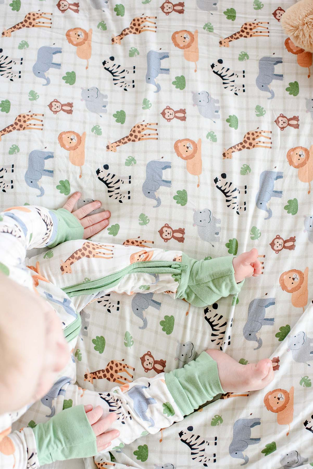 Overhead close-up of a baby sitting on a Caden Lane Wild Child bamboo blanket, showing the matching zip romper's green cuffs and side zipper.