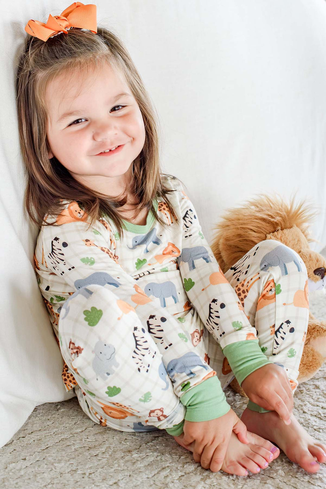Smiling girl sitting on a rug against a white couch, wearing a Caden Lane two-piece pajama set in a Wild Child print with green trim, paired with an orange hair bow and a lion plush.