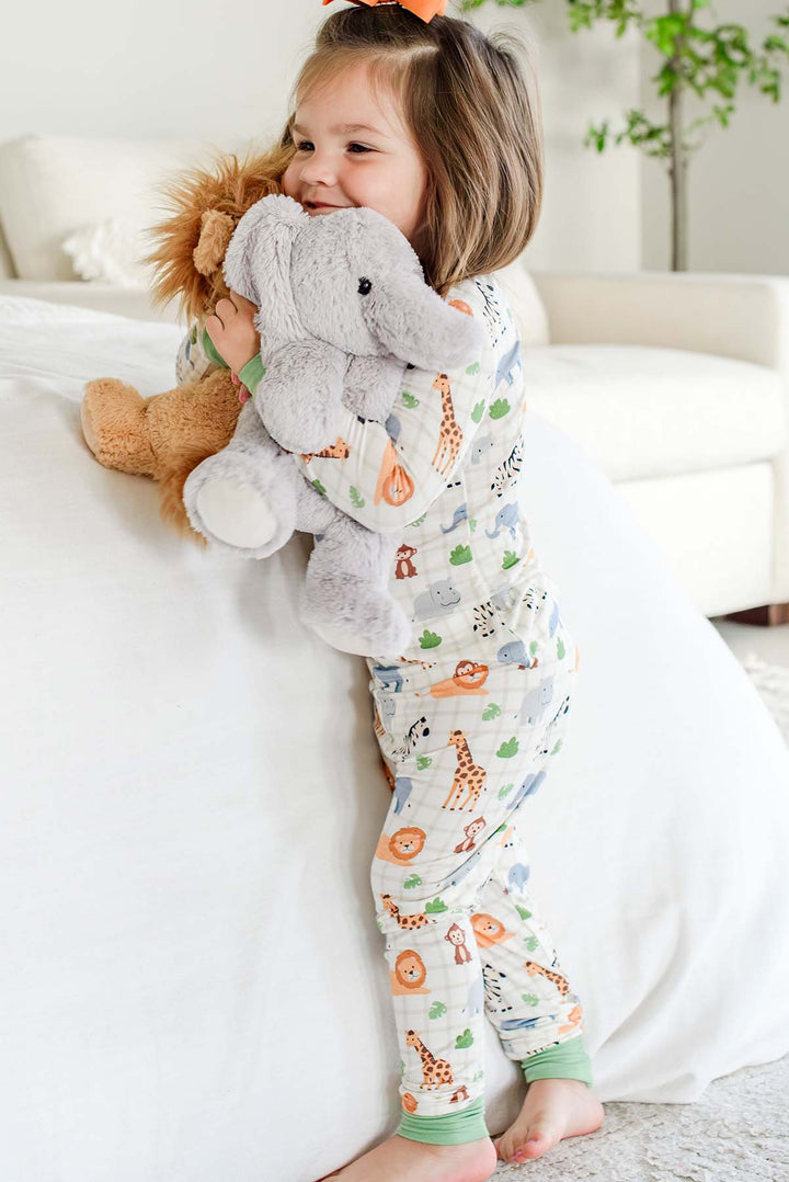 Girl leaning against a bed and hugging elephant and lion plush toys, wearing a Caden Lane two-piece pajama set in a Wild Child print featuring giraffes, lions and zebras.