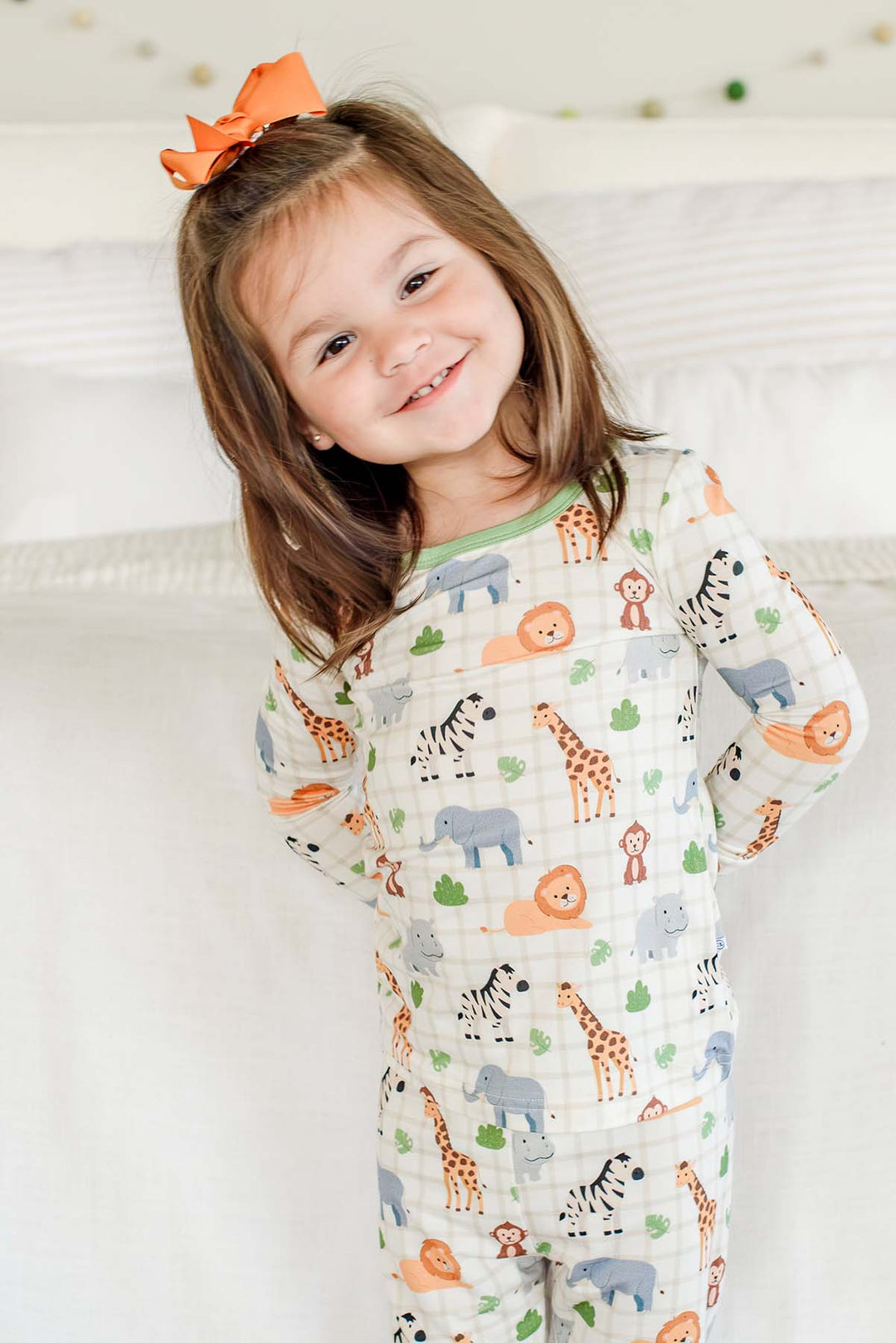 Smiling girl with her hands behind her back, wearing a Caden Lane Wild Child two-piece pajama set with a green neckline, paired with an orange hair bow.