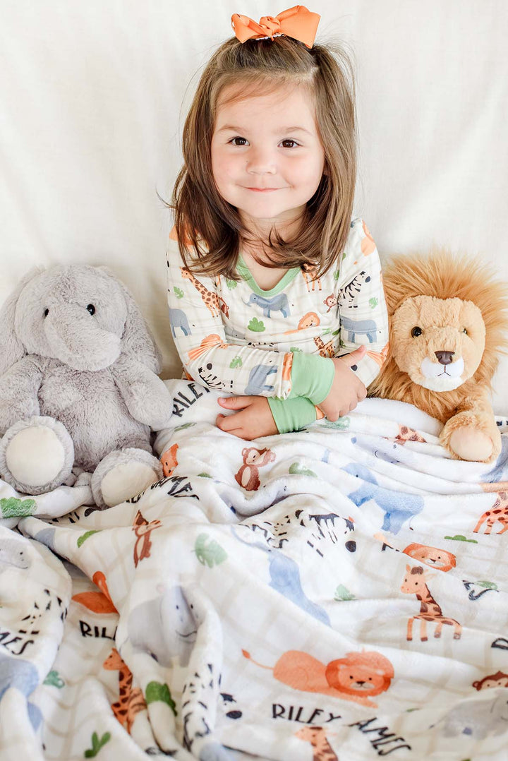 Smiling toddler girl sitting under a Caden Lane personalized plush blanket in a Wild Child print reading "Riley James," paired with matching Wild Child pajamas, an orange bow and elephant and lion plush toys.