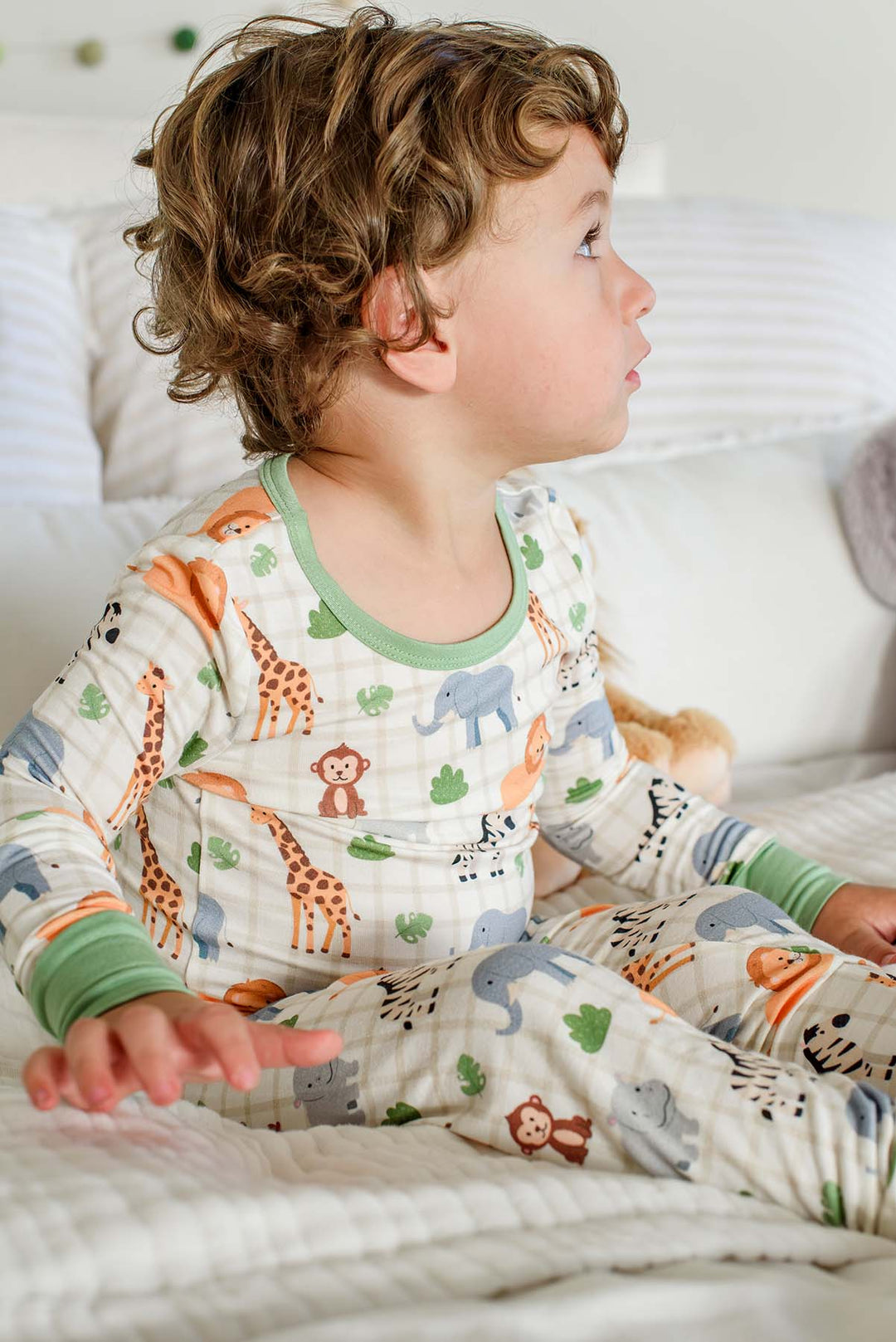 Side view of a curly-haired boy sitting on a bed and looking up, wearing a Caden Lane two-piece pajama set in a Wild Child print with green trim.