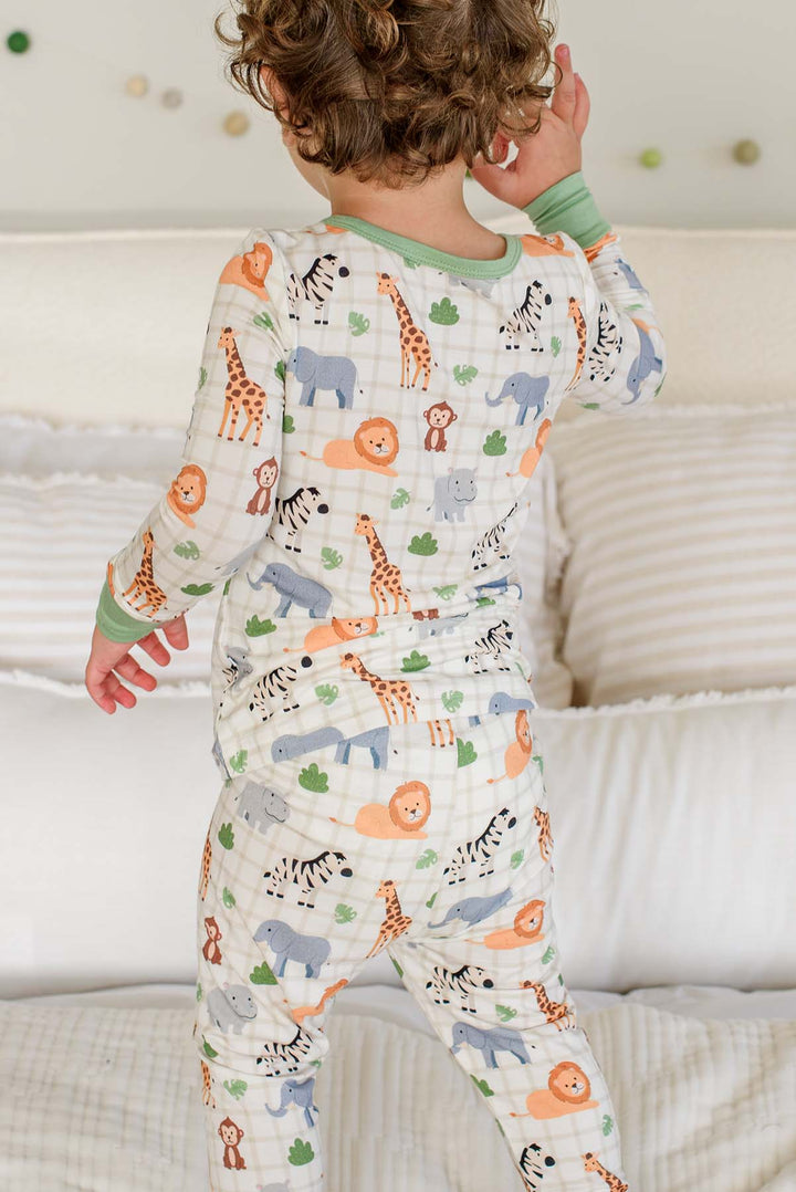 Back view of a curly-haired boy standing on a bed in a Caden Lane two-piece pajama set in a Wild Child print featuring giraffes, lions, zebras and elephants.
