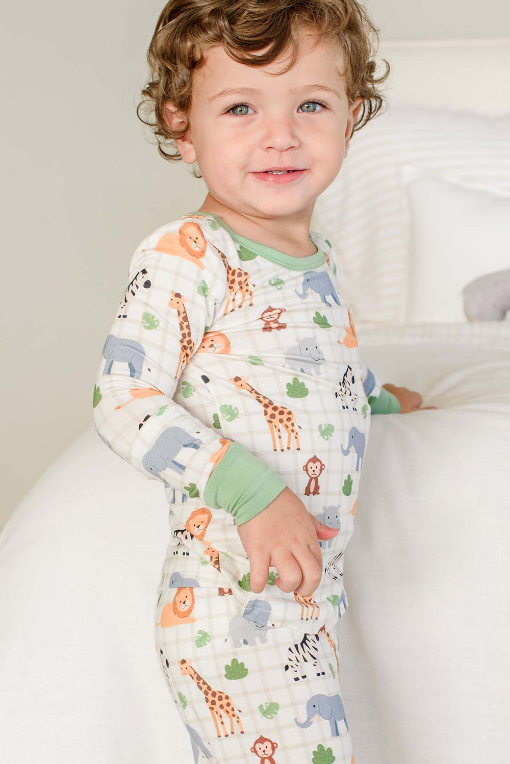 Smiling curly-haired boy standing beside a bed, wearing a Caden Lane two-piece pajama set in a Wild Child print featuring giraffes, monkeys, elephants and hippos.
