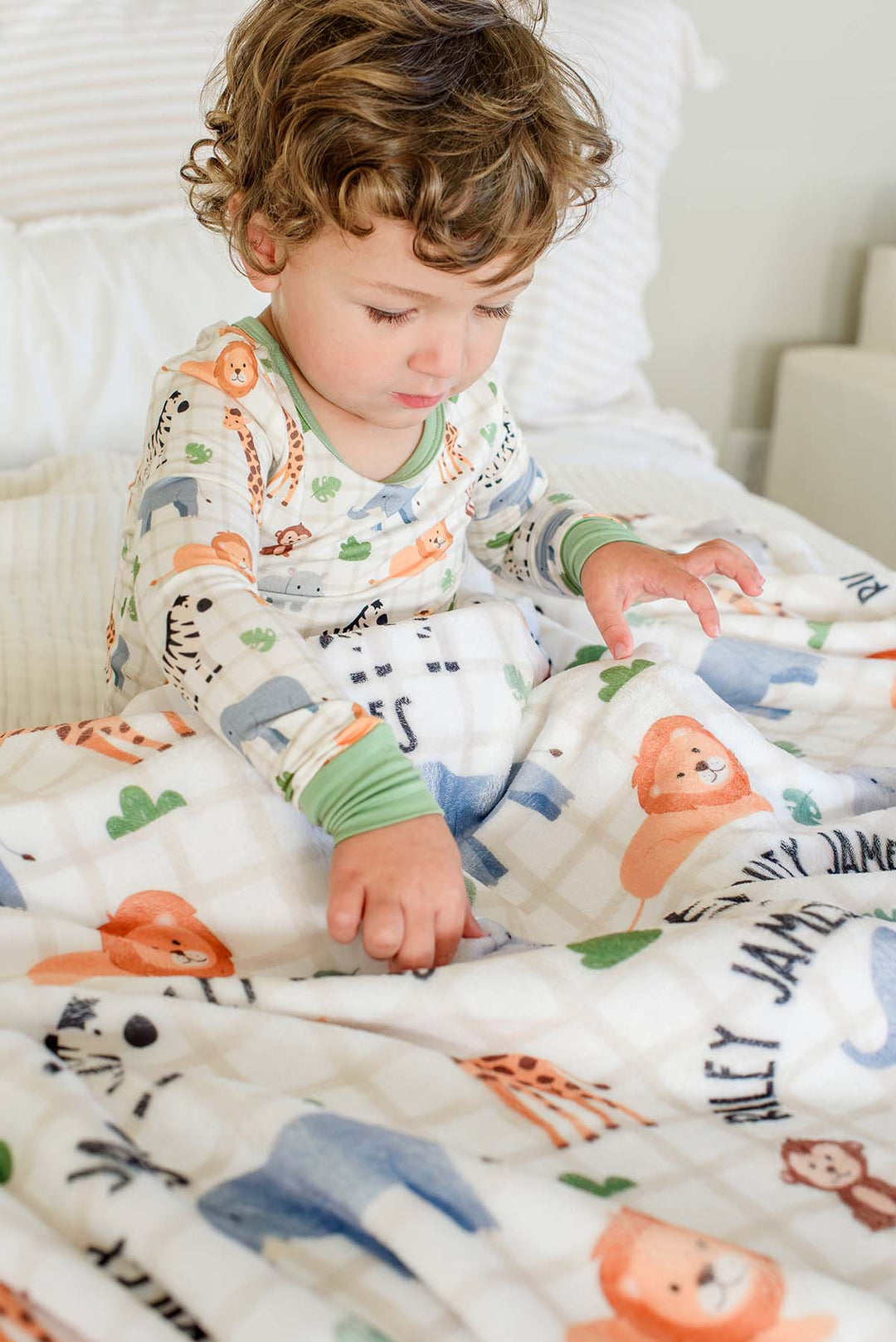 Curly-haired toddler boy sitting on a bed exploring a Caden Lane personalized plush blanket in a Wild Child print reading "Riley James," paired with matching green-trimmed pajamas.