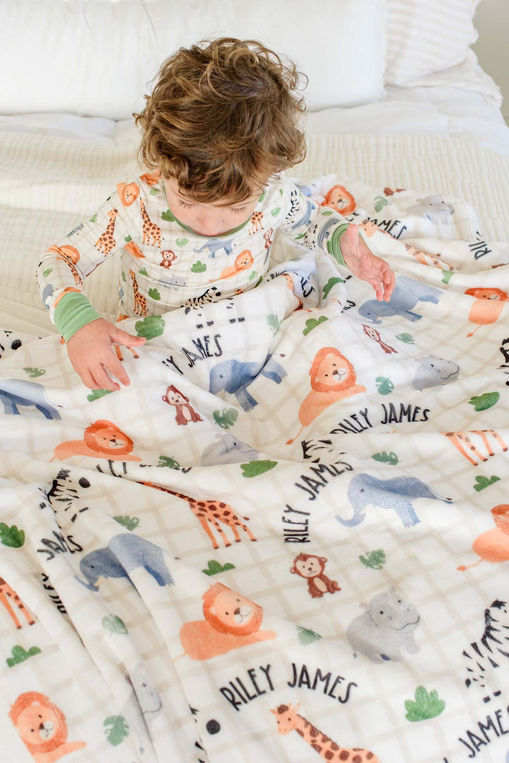 Overhead view of a toddler boy in matching Wild Child pajamas reaching across a Caden Lane personalized plush blanket reading "Riley James" spread over a bed.