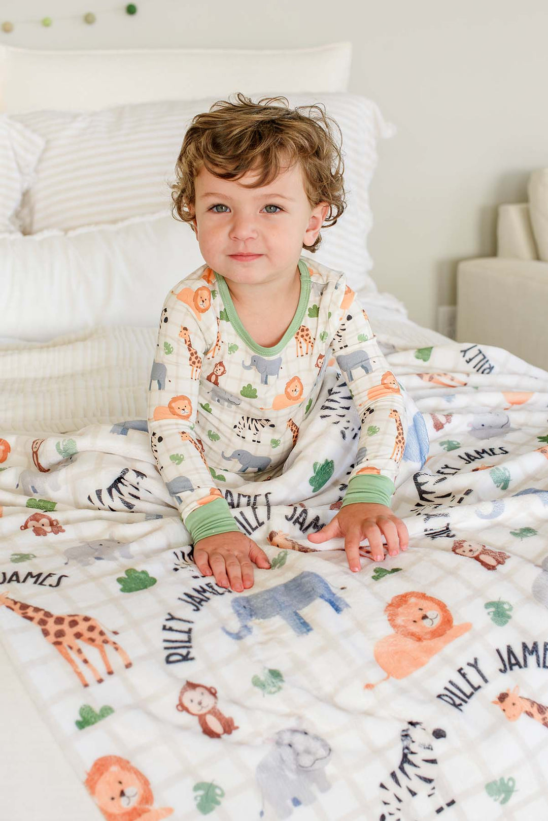 Toddler boy sitting on a bed with his hands resting on a Caden Lane personalized plush blanket in a Wild Child print reading "Riley James," paired with matching Wild Child pajamas.