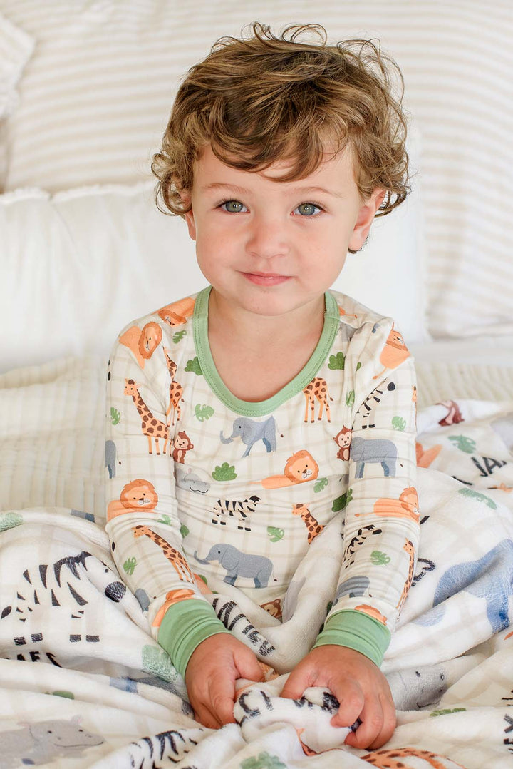 Curly-haired boy sitting on a bed in a Caden Lane Wild Child two-piece pajama set with green trim, holding a matching personalized plush blanket.