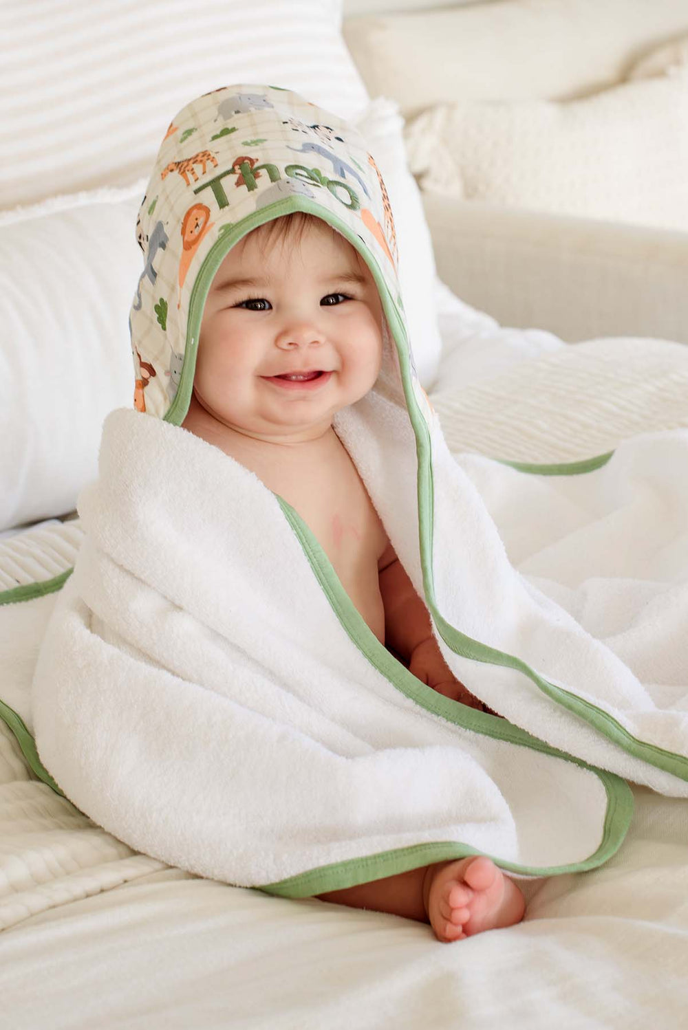 Smiling baby sitting on a bed wrapped in a Caden Lane white hooded towel with green trim and a Wild Child print hood embroidered with "Theo."