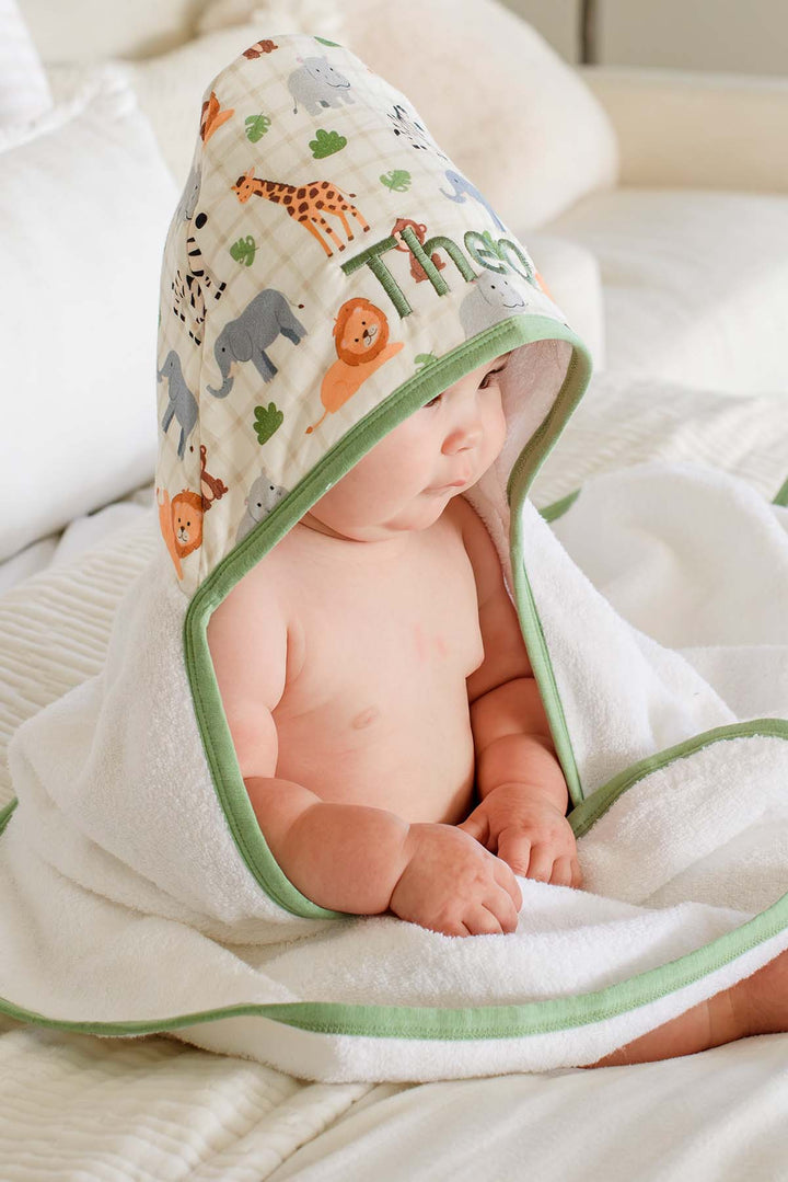 Side view of a baby looking away while wrapped in a Caden Lane hooded towel, showing the Wild Child print hood with giraffes, lions and elephants embroidered with "Theo."