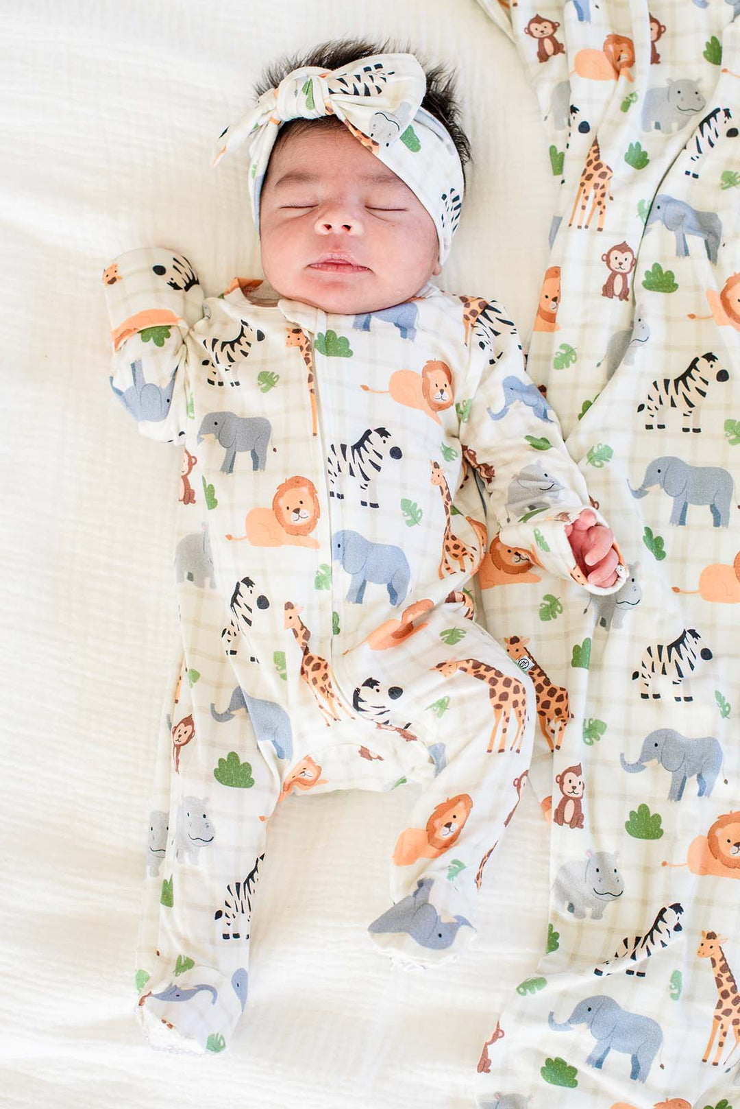 Sleeping newborn lying on a white bed in a Caden Lane zipper footie in a Wild Child print, paired with a matching knotted bow headwrap and blanket.