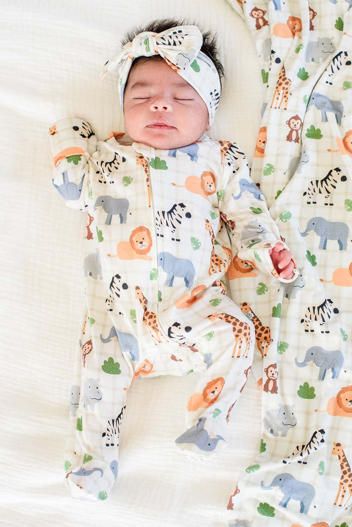 Sleeping newborn lying on a white bed in a Caden Lane zipper footie in a Wild Child print, paired with a matching knotted bow headwrap and blanket.