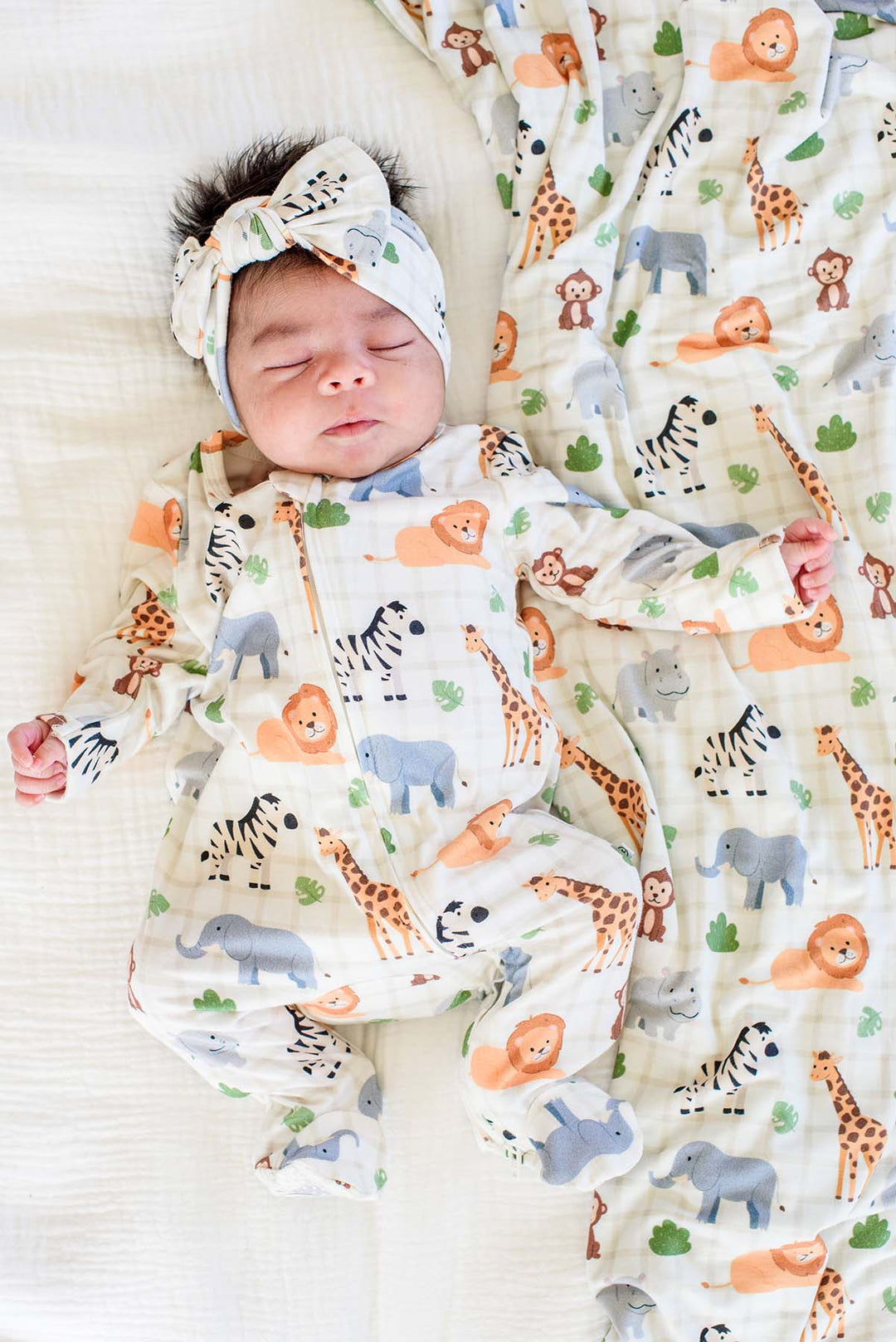 Overhead view of a sleeping newborn in a Caden Lane zipper footie in a Wild Child print, paired with a matching knotted bow headwrap and blanket.