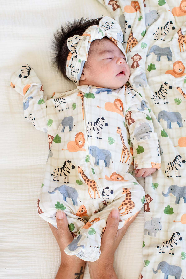 Parent's hands holding the feet of a sleeping newborn in a Caden Lane Wild Child zipper footie, showing the grip soles, paired with a matching headwrap and blanket.