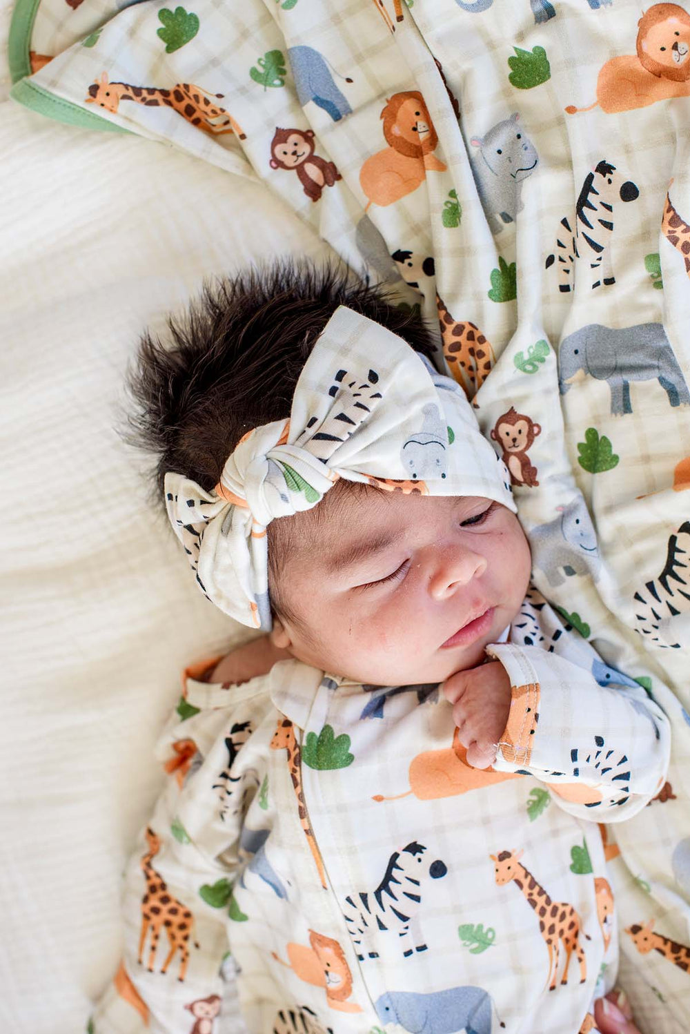 Overhead close-up of a sleeping newborn wearing a Caden Lane Wild Child knotted bow headwrap and matching footie, resting on a Wild Child blanket with green trim.