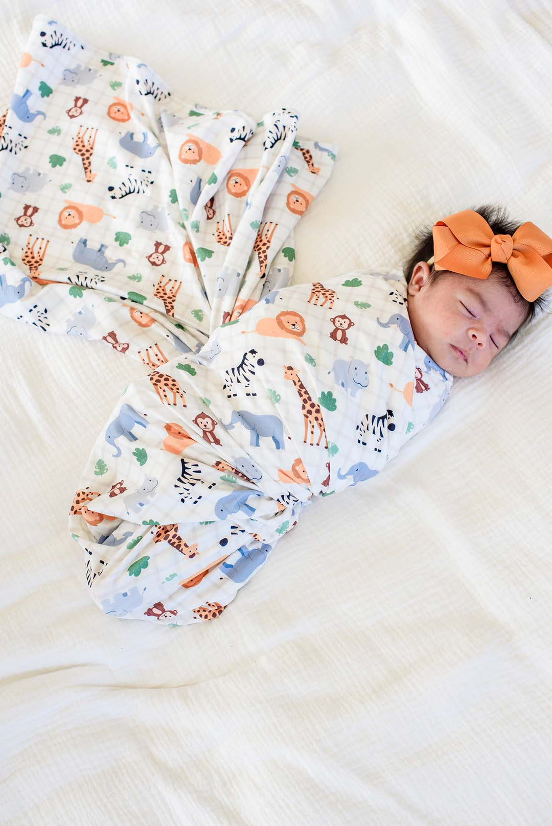 Overhead view of a sleeping newborn on a white bed, swaddled in a Caden Lane Wild Child swaddle blanket with the extra fabric fanned out, paired with an orange bow headband.
