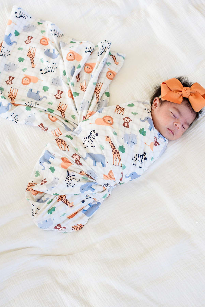 Overhead view of a sleeping newborn on a white bed, swaddled in a Caden Lane Wild Child swaddle blanket with the extra fabric fanned out, paired with an orange bow headband.