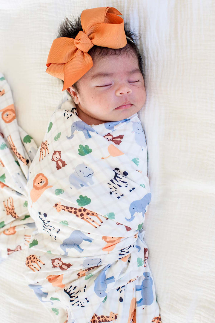Close-up of a sleeping newborn snugly swaddled in a Caden Lane Wild Child swaddle blanket with lions, zebras, elephants and giraffes, paired with an orange bow headband.