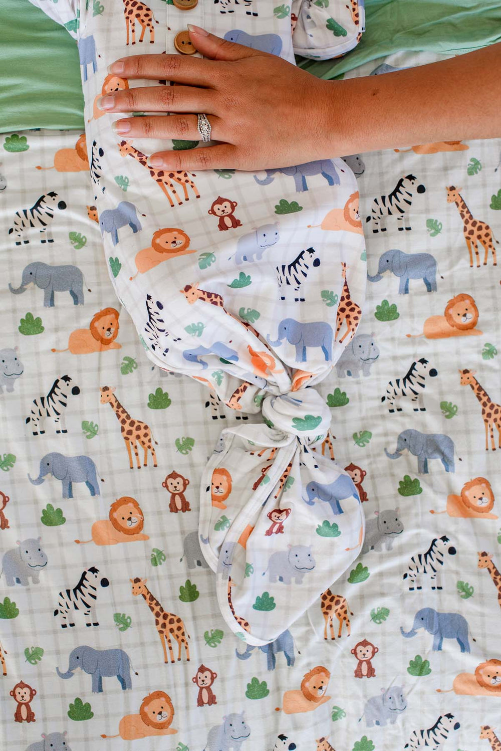 Close-up of a parent's hand resting on a newborn in a Caden Lane Wild Child knotted gown, showing the tied bottom against a matching safari animal blanket.