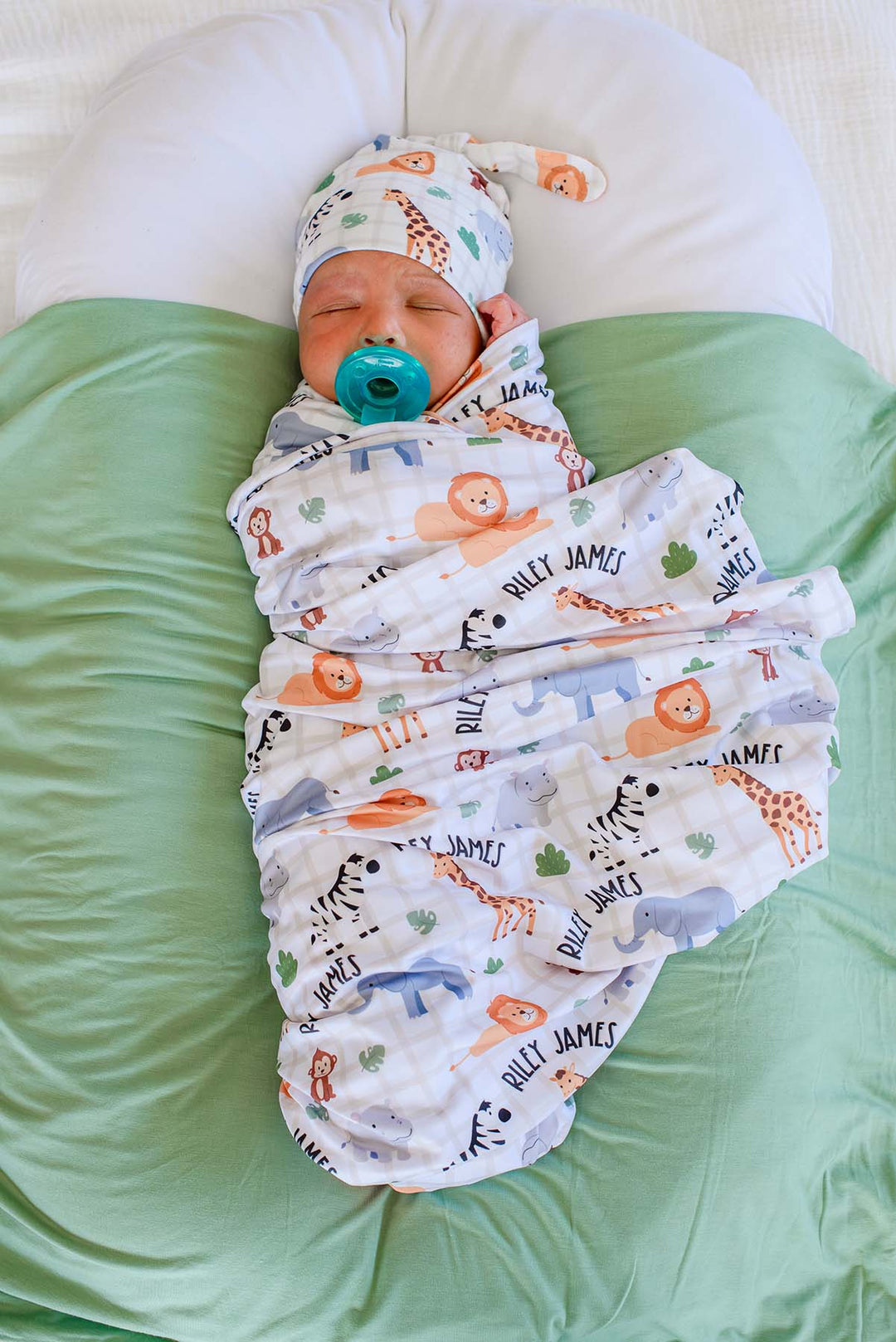 Overhead view of a sleeping newborn with a teal pacifier, swaddled in a Caden Lane personalized swaddle blanket in a Wild Child print reading "Riley James," paired with a matching knot hat on a sage green blanket.