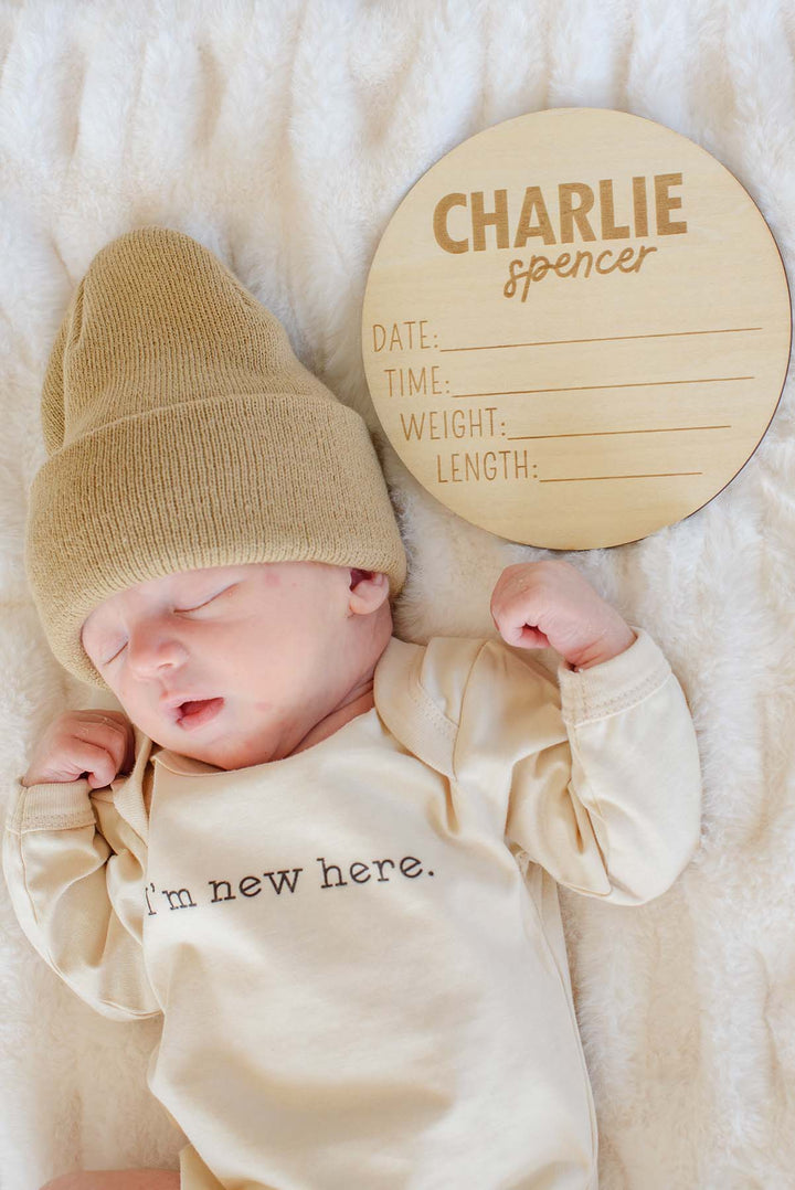 Newborn in a Caden Lane tan beanie and "I'm new here" cream bodysuit, sleeping on a faux fur blanket beside a wooden "Charlie Spencer" birth announcement plaque.