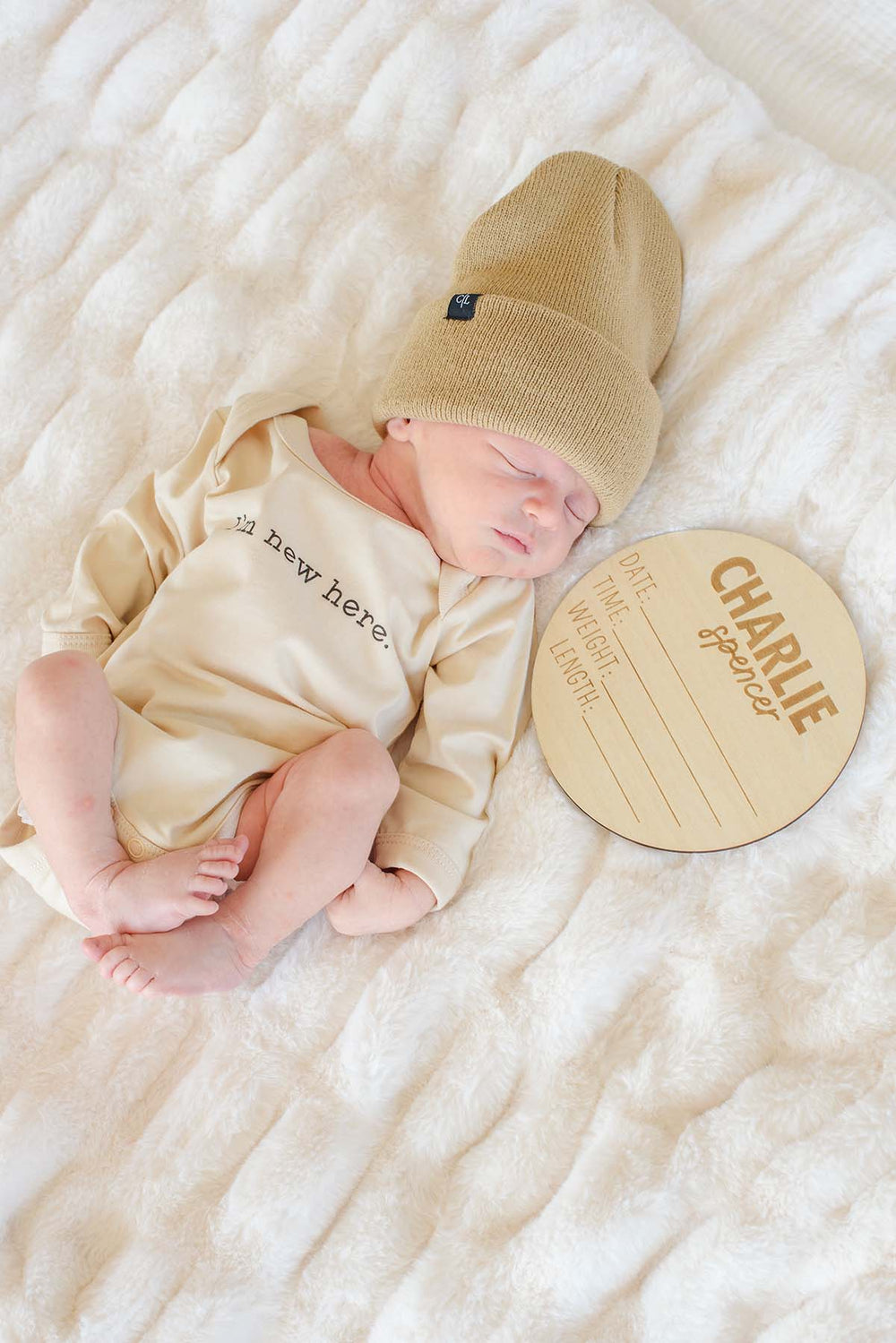 Sleeping newborn wearing a Caden Lane tan knit beanie with logo tag and cream "I'm new here" bodysuit, lying next to a personalized wooden "Charlie Spencer" name sign on a fuzzy white blanket.