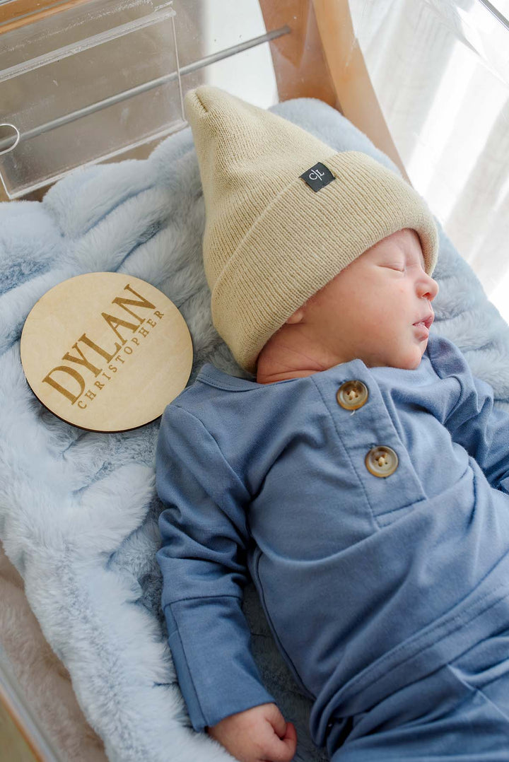 Sleeping newborn boy in a Caden Lane tan knit beanie with logo tag and blue button-shoulder outfit, lying in a hospital bassinet next to a wooden "Dylan Christopher" birth announcement sign.