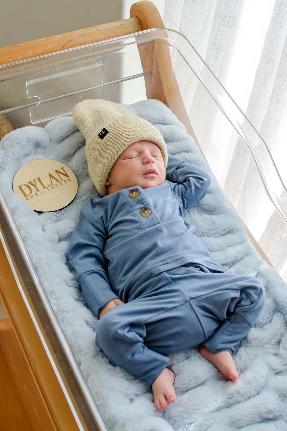 Newborn boy in a Caden Lane tan beanie and blue button-detail sleeper resting on blue faux fur in a hospital bassinet, beside a wooden "Dylan Christopher" name sign.