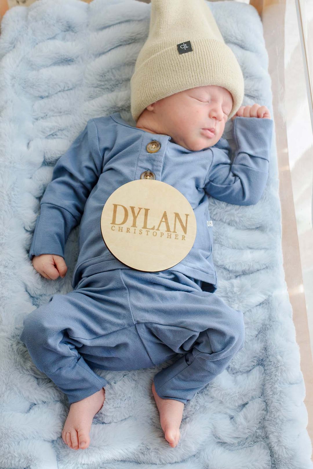 Close-up overhead view of a sleeping newborn in a Caden Lane tan knit beanie and blue button-shoulder outfit, with a Caden Lane wooden "Dylan Christopher" sign resting on his chest.