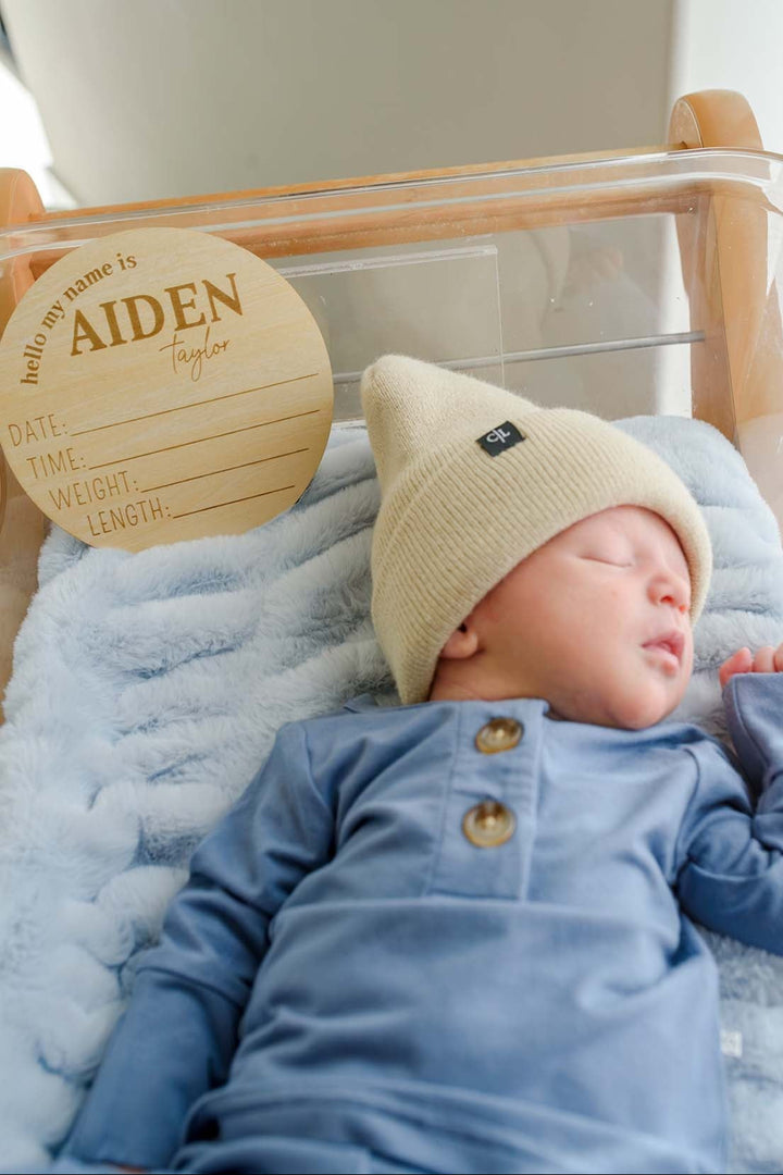 Sleeping newborn boy wearing a Caden Lane blue button-shoulder outfit and tan knit beanie with Caden Lane logo tag, lying in a hospital bassinet beside a wooden "hello my name is Aiden Taylor" birth announcement sign.