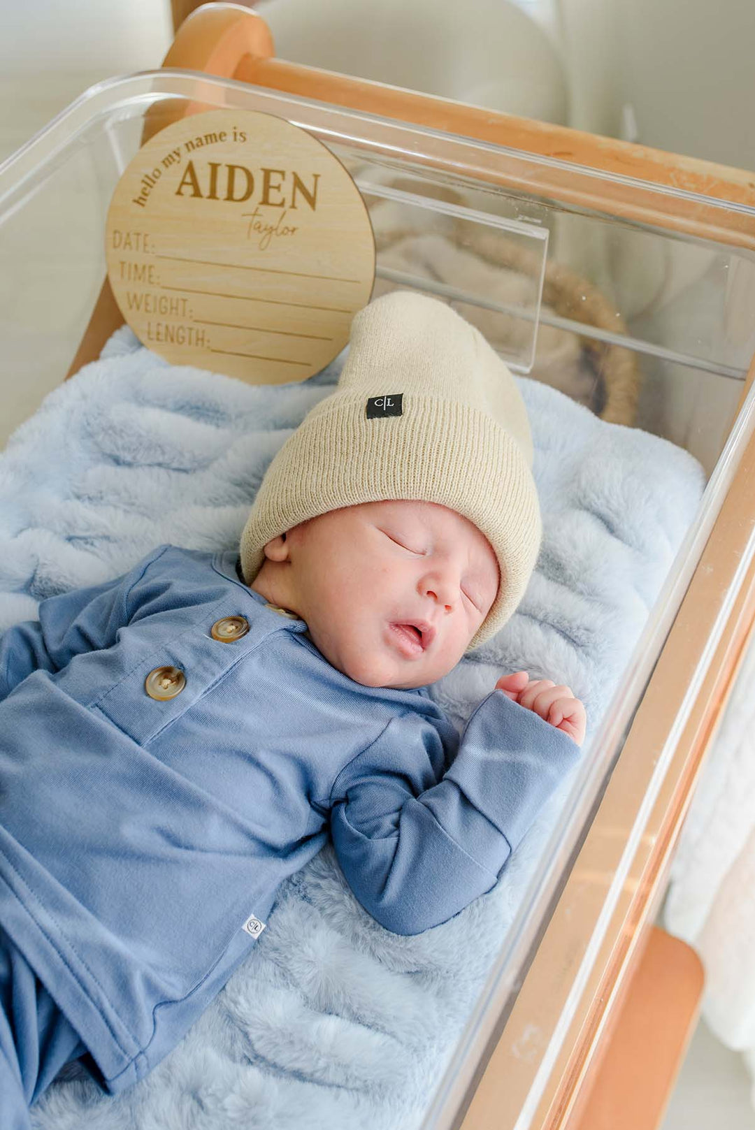Newborn boy in a Caden Lane blue button-detail sleeper resting on blue faux fur in a hospital bassinet, next to a Caden Lane engraved wood sign reading "hello my name is Aiden Taylor."