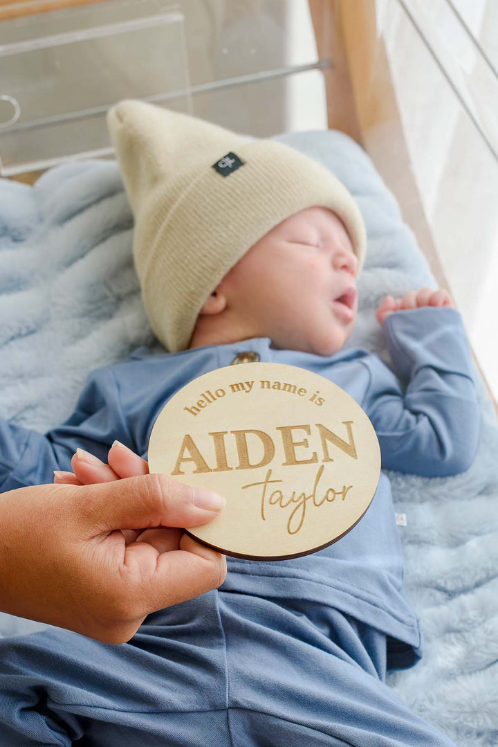 A hand holds up a Caden Lane wooden "hello my name is Aiden Taylor" sign in front of a sleeping newborn boy wearing a Caden Lane tan knit beanie with logo tag and blue button-shoulder outfit in a hospital bassinet.