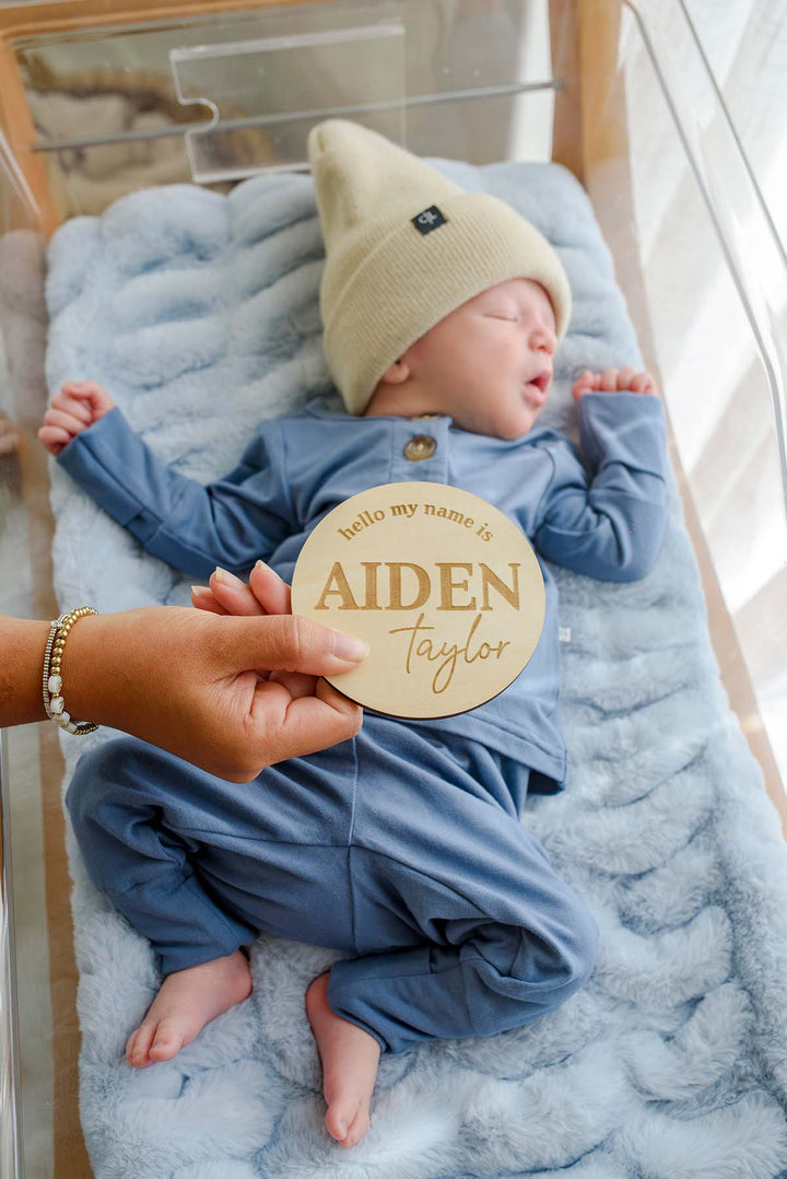 Overhead view of a hand holding a Caden Lane engraved wood name sign reading "hello my name is Aiden Taylor" over a sleeping newborn in a Caden Lane tan beanie and blue sleeper.