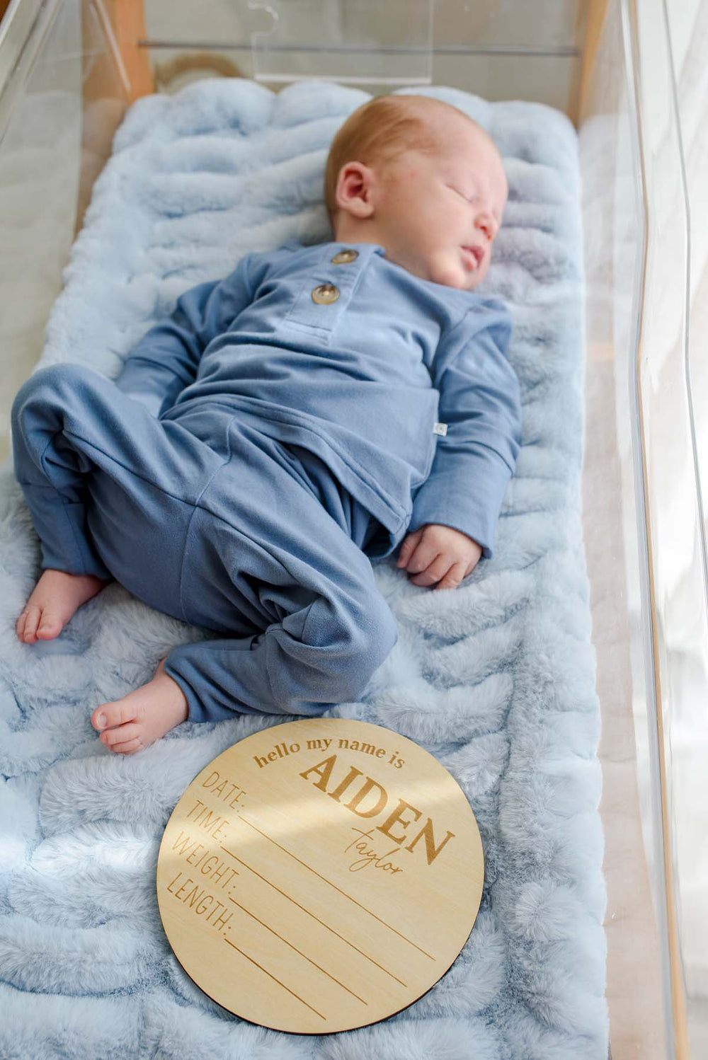 Close-up of a sleeping newborn in a Caden Lane tan knit beanie with logo tag and blue button-shoulder outfit, positioned beside a wooden "hello my name is Aiden Taylor" birth announcement plaque in a hospital bassinet.