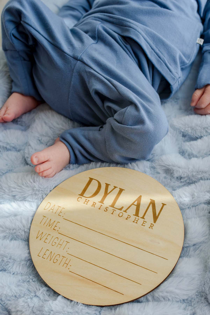 Close-up of a newborn's feet in a Caden Lane blue knit outfit, lying beside a Caden Lane personalized wooden birth announcement sign reading "Dylan Christopher" with blank date, time, weight, and length lines on a blue faux fur blanket.