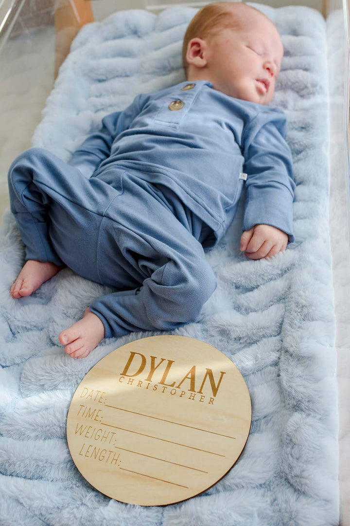Sleeping newborn boy in a Caden Lane blue button-detail sleeper, lying in a bassinet on blue faux fur next to a wooden "Dylan Christopher" name sign.