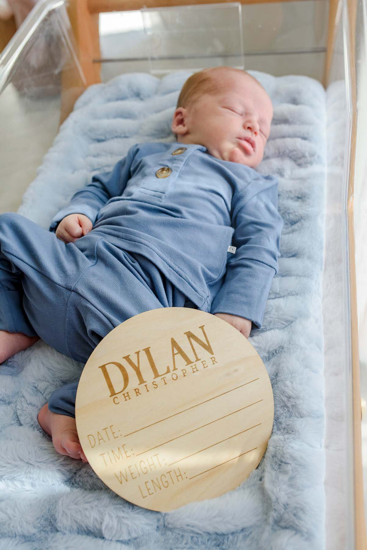 Newborn boy in a Caden Lane dusty blue sleeper resting in a hospital bassinet, with a Caden Lane engraved wood birth announcement plaque reading "Dylan Christopher" propped against his feet.