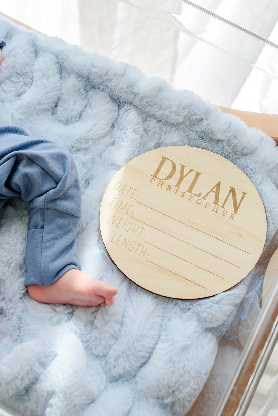 Close-up of a newborn's foot in a Caden Lane blue sleeper beside a wooden "Dylan Christopher" birth announcement sign on a blue faux fur blanket in a hospital bassinet.