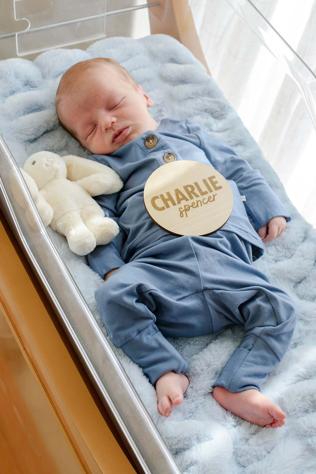 Newborn boy in a Caden Lane blue button-detail outfit sleeping in a hospital bassinet, with a Caden Lane wooden "Charlie Spencer" name sign placed on his chest and a bunny stuffed animal beside him.