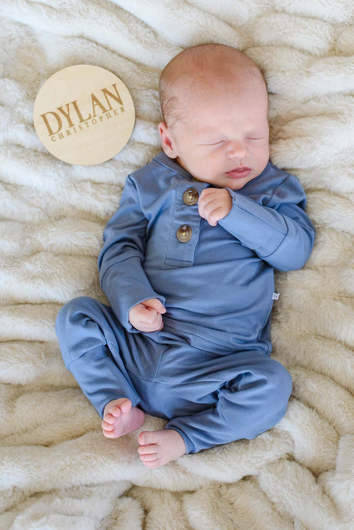 Newborn boy in a Caden Lane blue button-shoulder sleeper lying on a cream faux fur blanket beside a wooden "Dylan Christopher" birth announcement plaque.