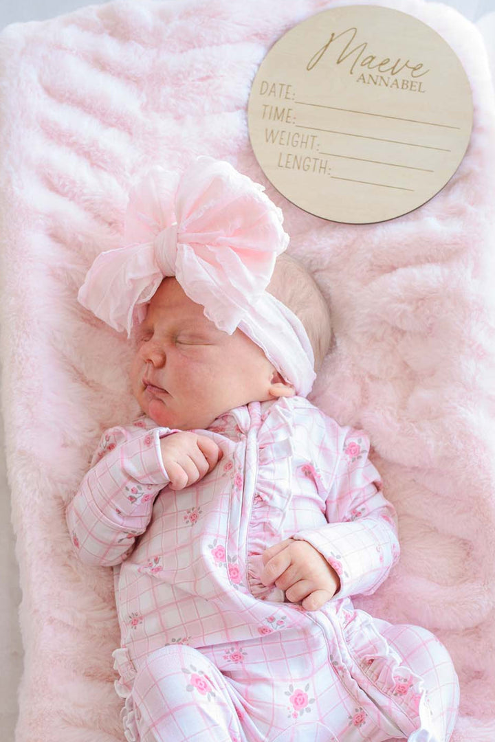 Sleeping newborn girl in a Caden Lane pink floral windowpane sleeper gown with a pink bow headband, lying beside a personalized wooden birth announcement sign reading "Maeve Annabel" with date, time, weight, and length fields.