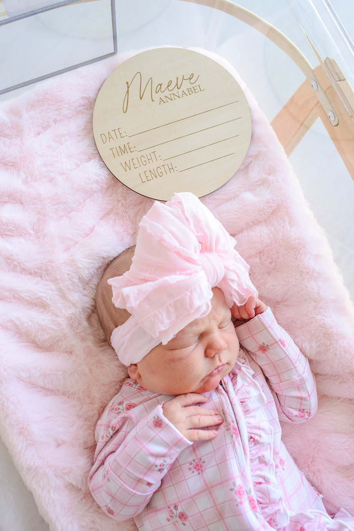 Close-up of a newborn wearing Caden Lane's pink floral windowpane footie pajama with ruffle trim, sleeping next to a Caden Lane engraved wood birth announcement sign in a bassinet lined with pink faux fur.