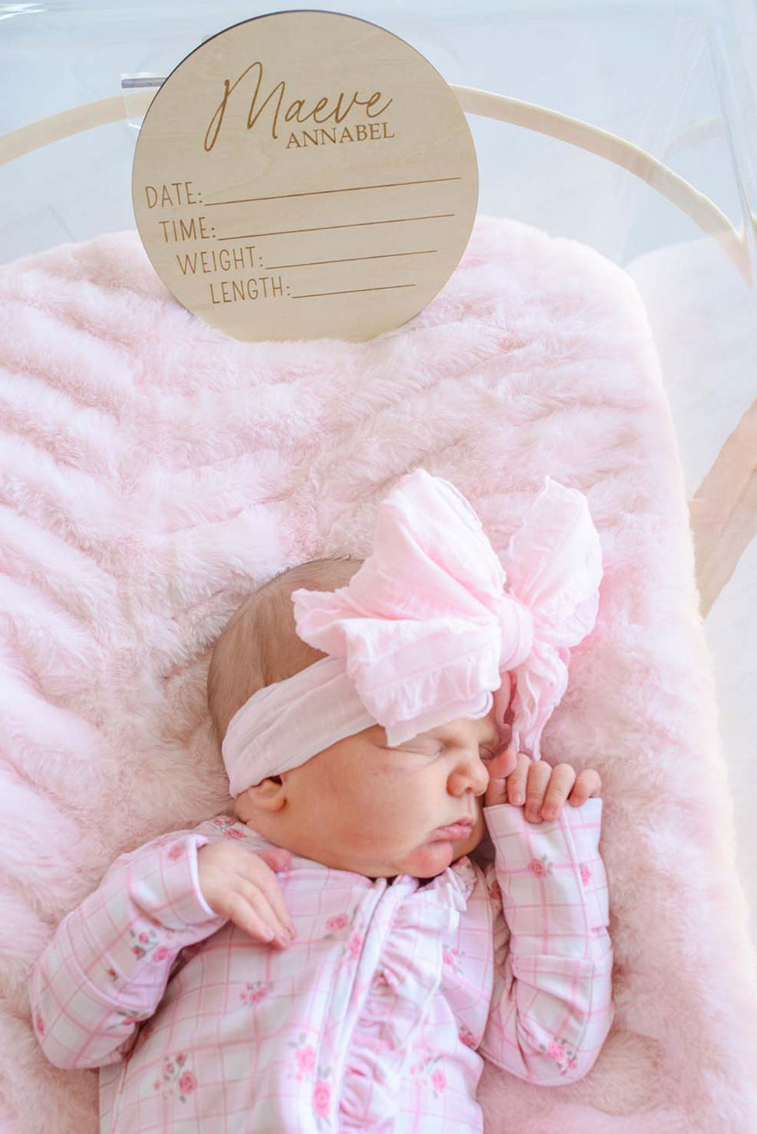 Overhead view of a newborn baby in a Caden Lane pink floral gown and bow headband, resting beneath a wooden "Maeve Annabel" name sign with blank date, time, weight, and length lines for a hospital photo.